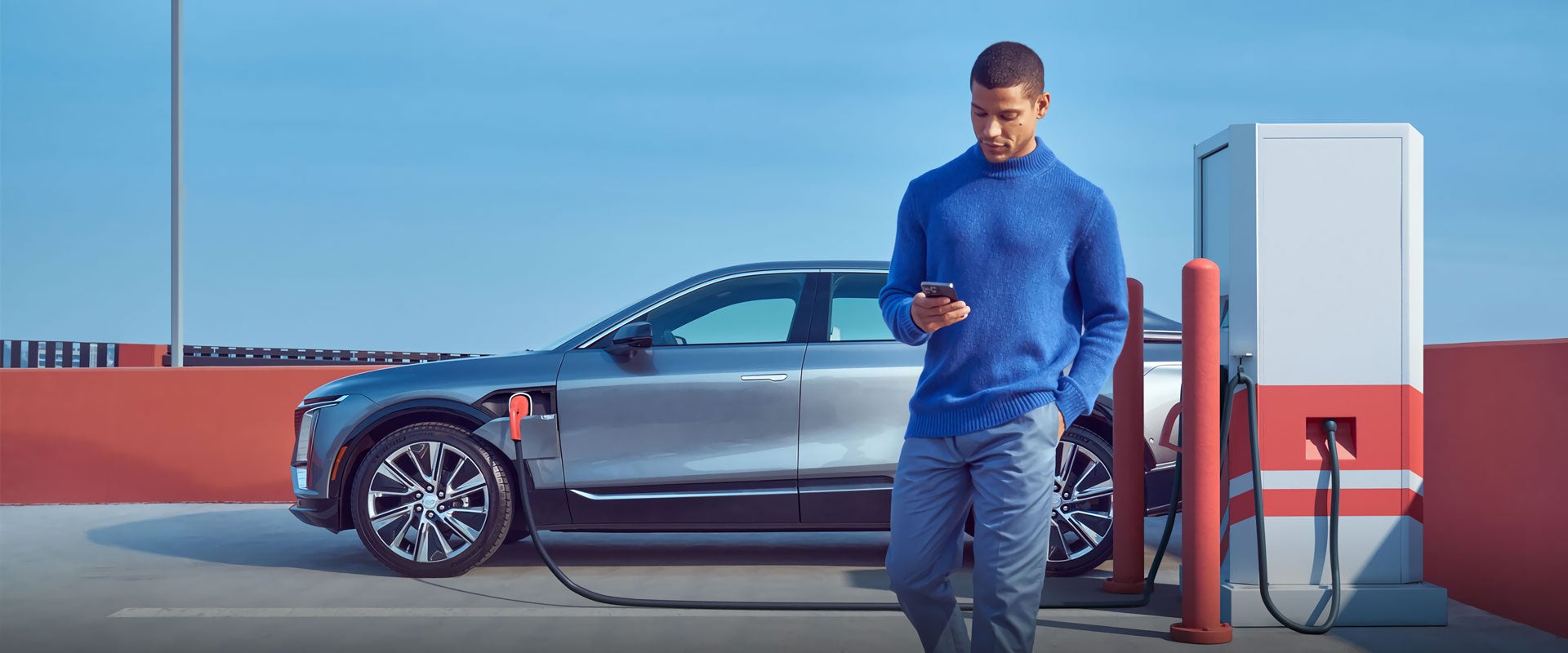 Photo of a person looking down at their phone with an electric Cadillac vehicle charging in the background